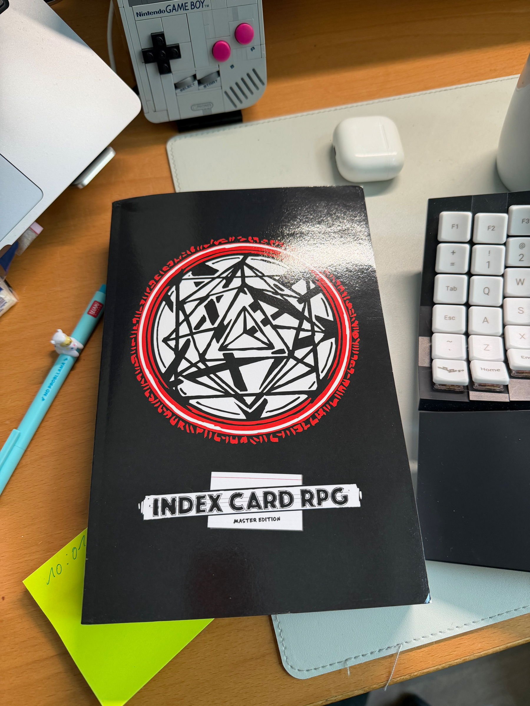 A phot of the Index Card RPG Master Edition placed on a desk. Other visible items:&10;&10;- post-it-notes&10;- a No Probllama-pen&10;- a Lego Gameboy&10;- a Moergo Glove80&10;- a case of wireless headphones&10;- part of a laptop